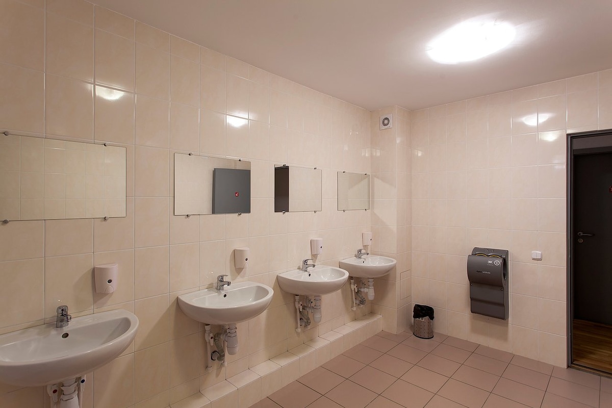 A well-lit communal restroom features three sinks equipped with mirrors above each. Soft lighting enhances the clean, tiled walls, and a paper towel dispenser is visible on the side. A waste bin is positioned near the sinks for convenience.