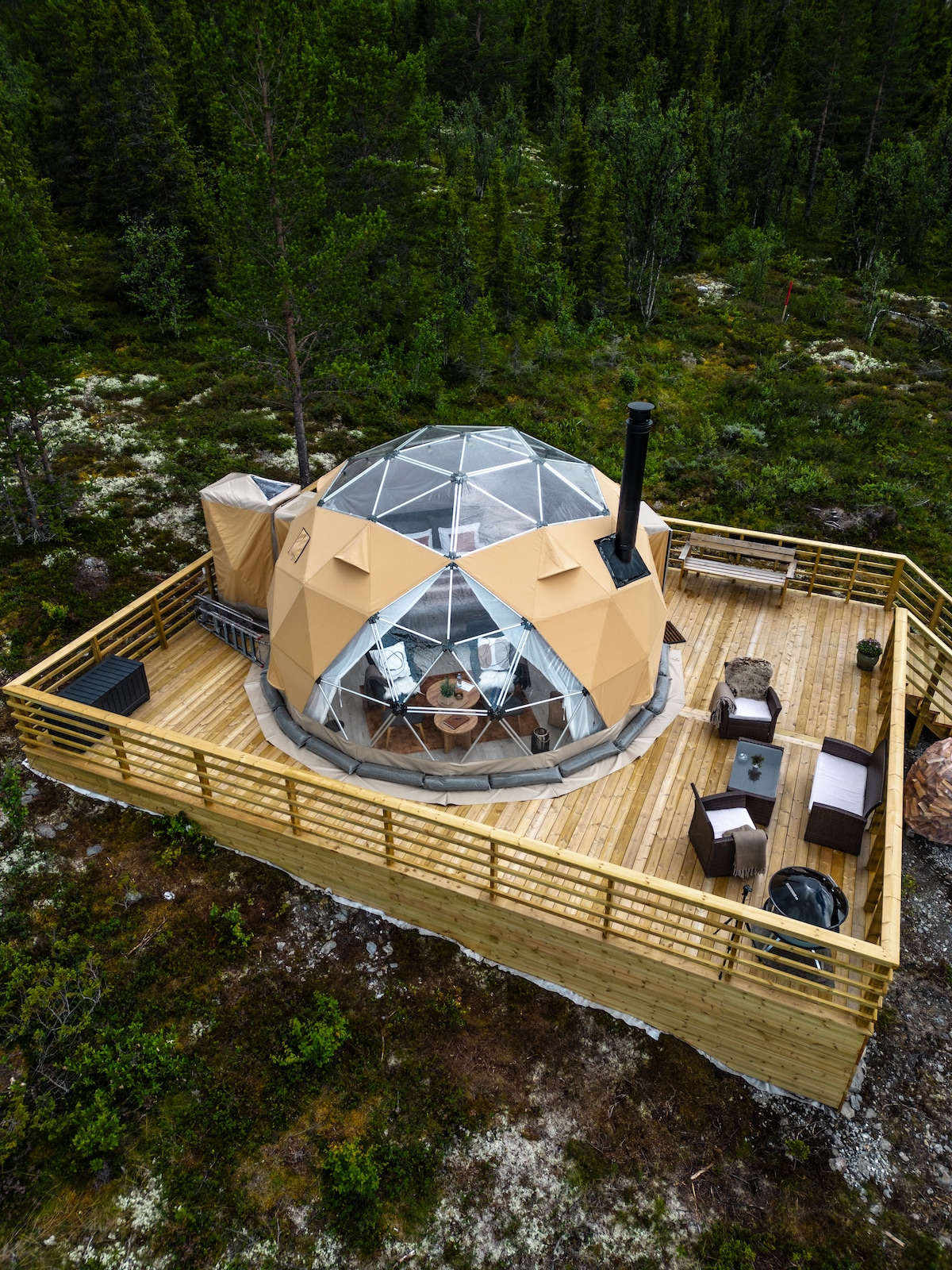 An aerial view of a geodesic dome set within a natural landscape. Surrounded by trees, the dome features a wooden deck with comfortable seating and a fire pit. The structure is topped with a glass panel, allowing for views of the sky above.