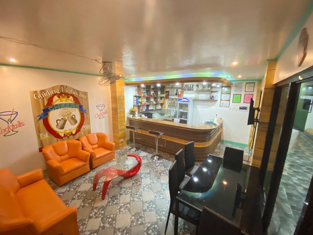 A communal lounge area is presented with vibrant orange chairs and a sleek red coffee table. A well-stocked bar is equipped with shelves of beverages and seating arrangements. The patterned flooring complements the aesthetic, while wall decorations enhance the inviting ambiance.
