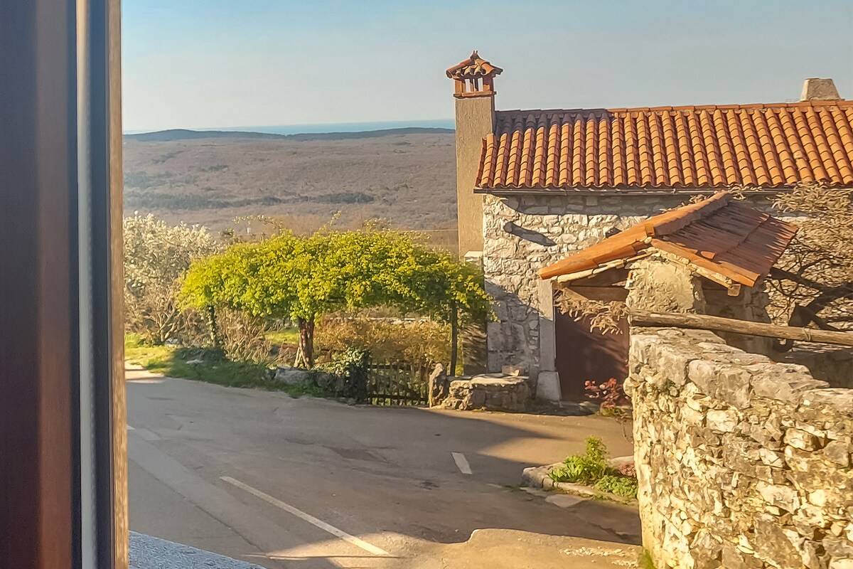 A view from the window captures a rustic stone house featuring a terracotta roof and a prominent chimney. Lush green trees frame the scene, overlooking a winding road that leads to expansive natural landscapes in the distance.