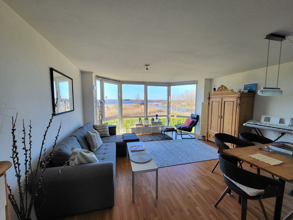 A spacious living area is filled with natural light, featuring a comfortable sofa and a dining table. Large windows offer views of the surrounding landscape. A wooden cabinet stands against the wall, while a small coffee table is placed in front of the sofa.