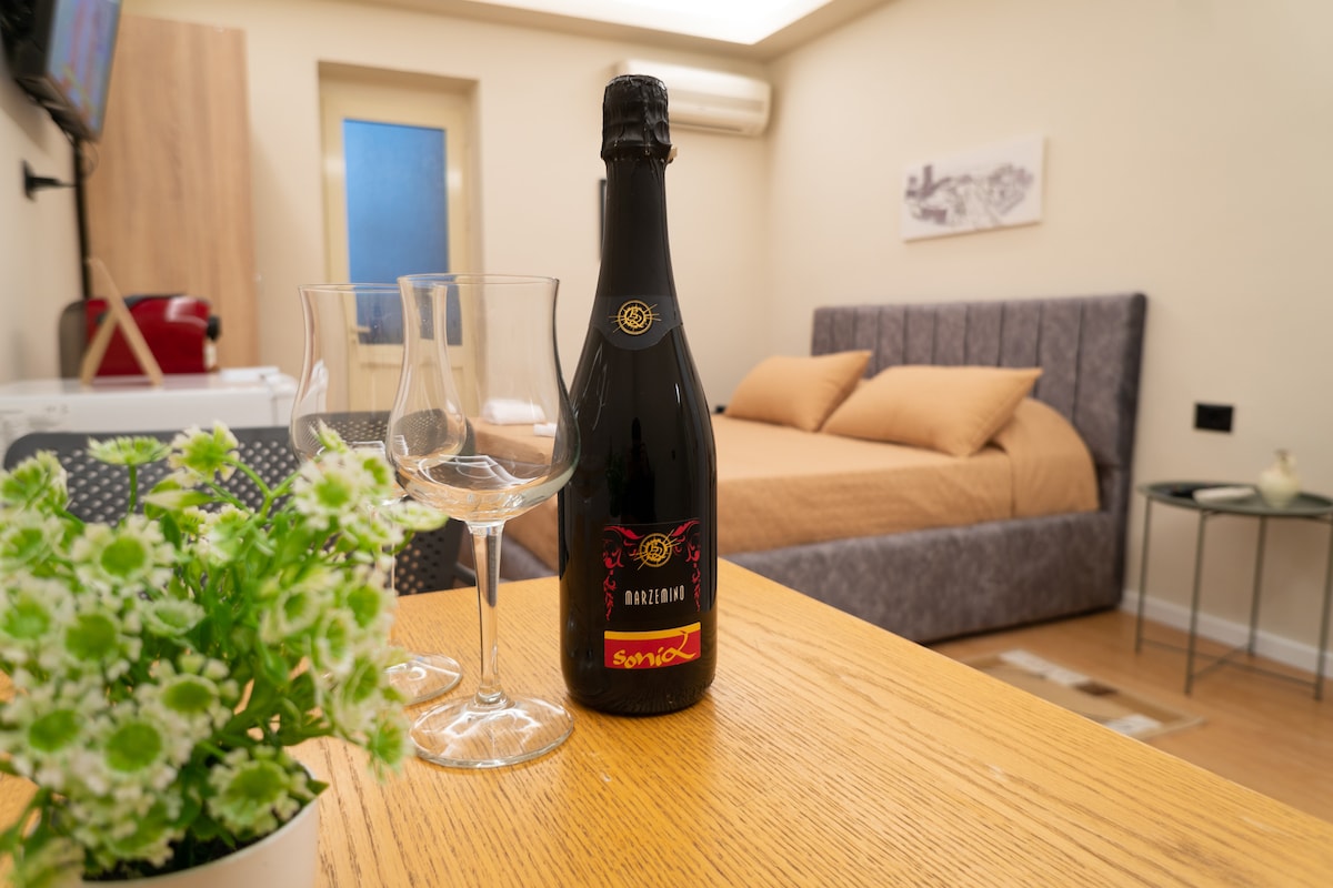 A bottle of sparkling wine is presented on a wooden table, accompanied by two clear glasses. In the background, a cozy sleeping area features a plush bed with neutral-colored bedding, while a small decorative plant adds a touch of greenery.