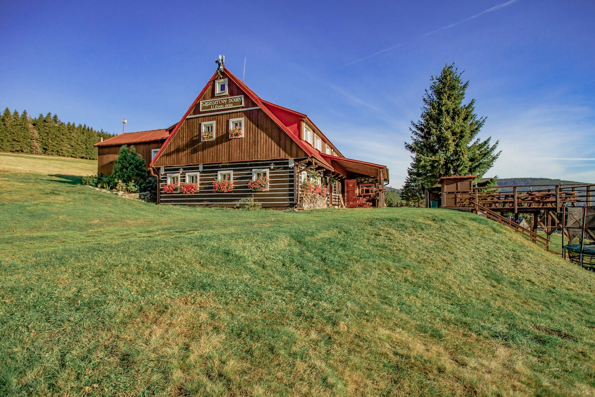The spacious cabin features a red-roof design with wooden siding, surrounded by green grassy areas. Large windows showcase vibrant floral decorations, and a wooden deck extends from the structure, offering views of the natural landscape beyond.
