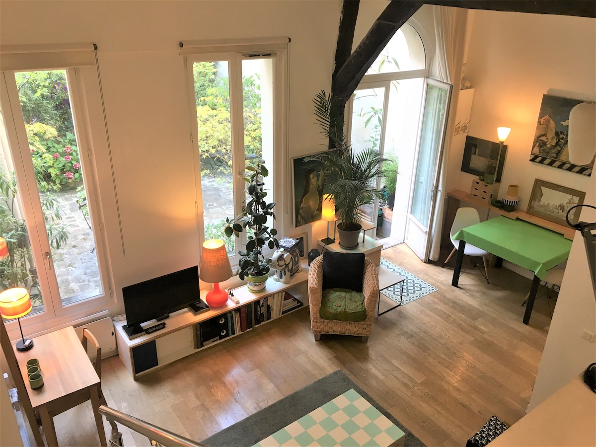 An aerial view captures a spacious living area featuring large windows that open to a serene terrace. A cozy seating arrangement includes a wicker chair and a green table. A wooden shelf holds a television and decorative items, while plants add a touch of nature.