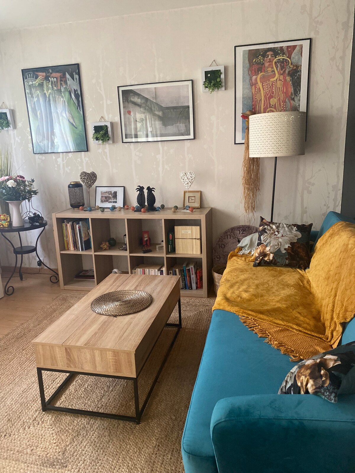 A cozy living space features a bright blue sofa adorned with a mustard throw. A wooden coffee table sits at the center, and a shelf displays an array of decorative items and books. Wall art adds character, and plants are positioned on shelves.