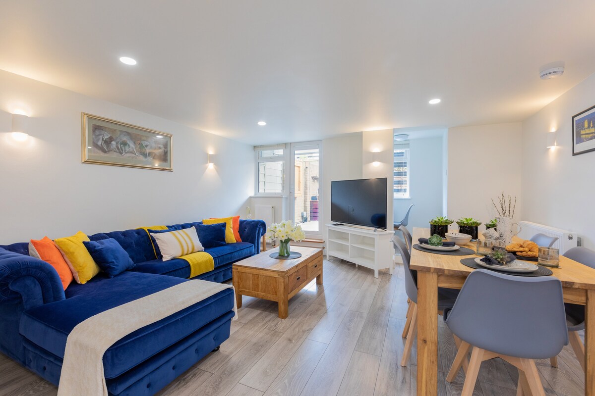 A spacious living and dining area is depicted, featuring a blue sectional sofa adorned with colorful cushions. A wooden coffee table anchors the room, complemented by a modern dining set. Large windows let in natural light, with a view of a private courtyard garden in the background.