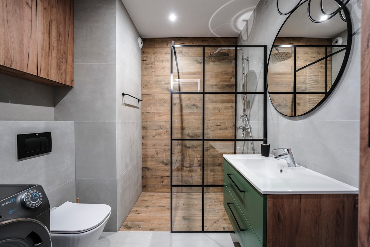 A modern bathroom features a spacious shower enclosed by glass panels, complemented by wooden accents on the walls. A large round mirror is mounted above the sink, which is positioned on a stylish vanity. The area includes a toilet and an electric washing machine.