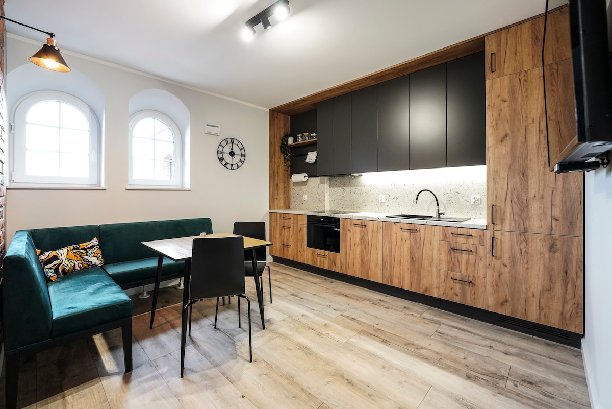 The modern kitchen area features a mix of wood and dark cabinetry, with a stone countertop and stainless steel appliances. A dining table is positioned beside a cozy green sofa adorned with a colorful cushion. Natural light filters through arched windows, enhancing the inviting ambiance.