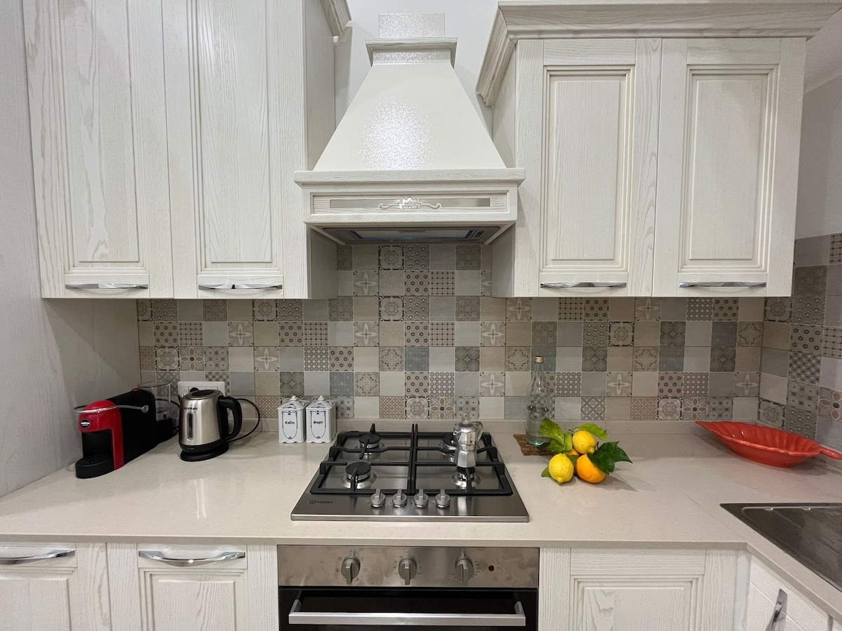 The kitchen features white cabinetry and a modern stove with a stainless steel hood. A patterned backsplash adds visual interest. Countertop appliances include a coffee machine and a kettle, complemented by a bowl of fresh fruit and decorative items on display.