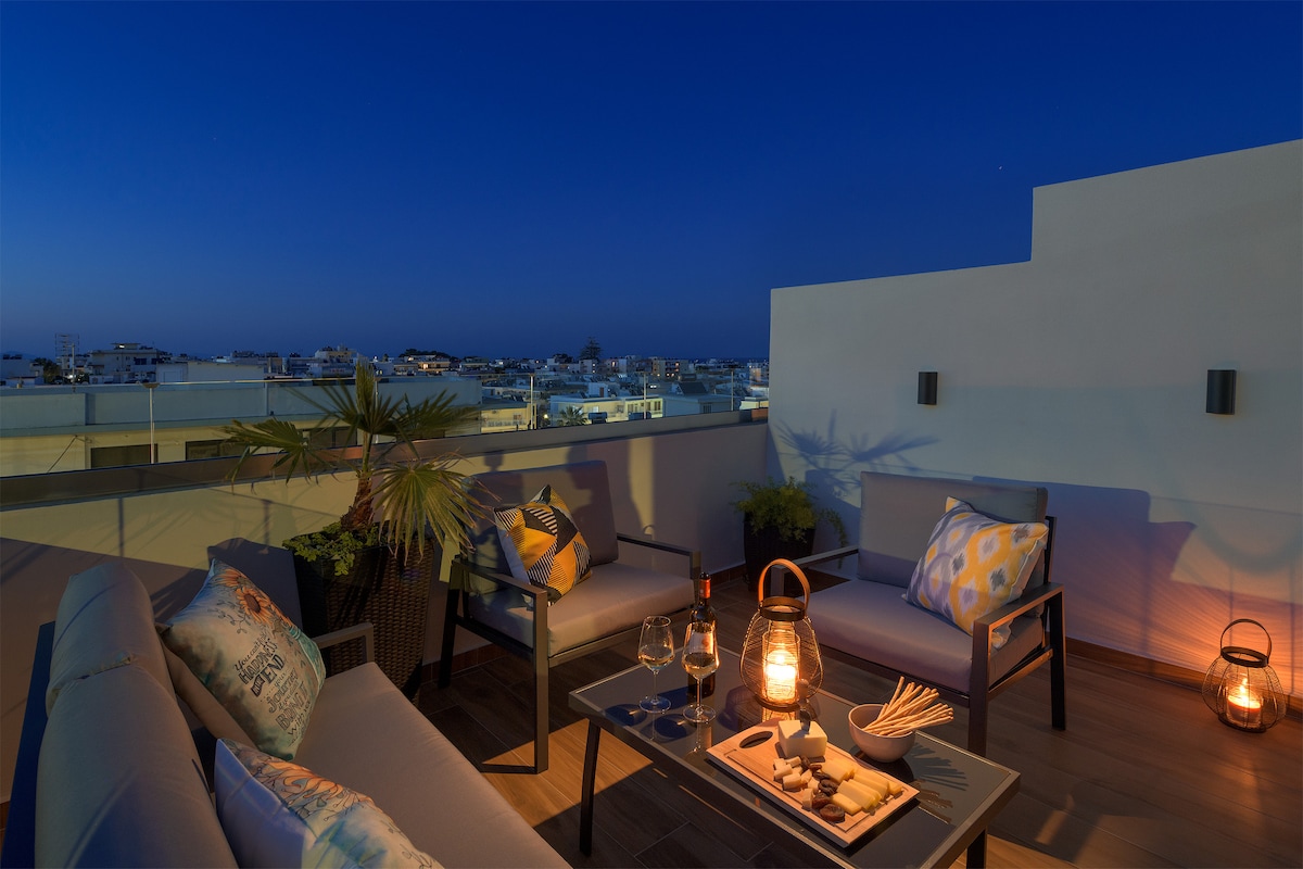 A spacious terrace is shown, furnished with comfortable seating and a low table. The scene is illuminated by lanterns, creating a relaxed ambiance. The backdrop displays a panoramic view of the surrounding area against a twilight sky.