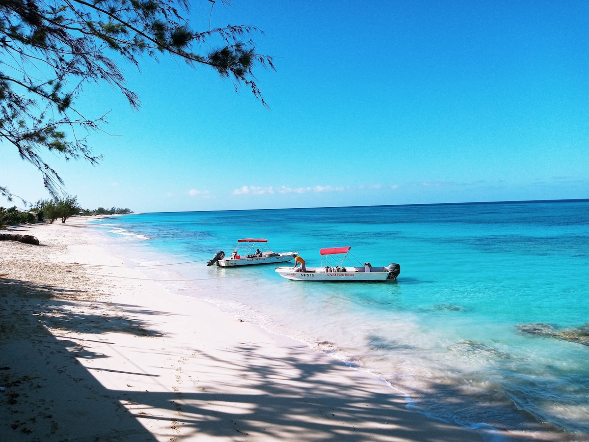 A tranquil beach scene is depicted, featuring two small boats anchored close to the shoreline. The sandy beach gently curves into crystal-clear turquoise waters, under a bright blue sky dotted with a few clouds. The shade from nearby trees adds to the serene atmosphere.