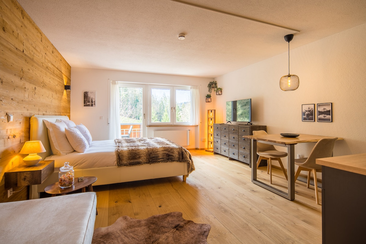 A spacious studio apartment is presented in a chalet style. A large bed with a soft throw is positioned beside a wooden feature wall. A dining table with light-colored chairs sits adjacent to a modern TV. Natural light floods in through the large windows.
