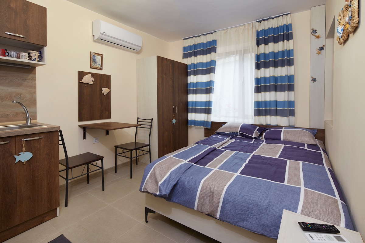 A bright studio space features a spacious bed layered with patterned bedding. A kitchenette is visible, accompanied by two dining chairs at a compact table. The window is adorned with striped curtains, allowing natural light to enter, while the walls are decorated with subtle maritime accents.