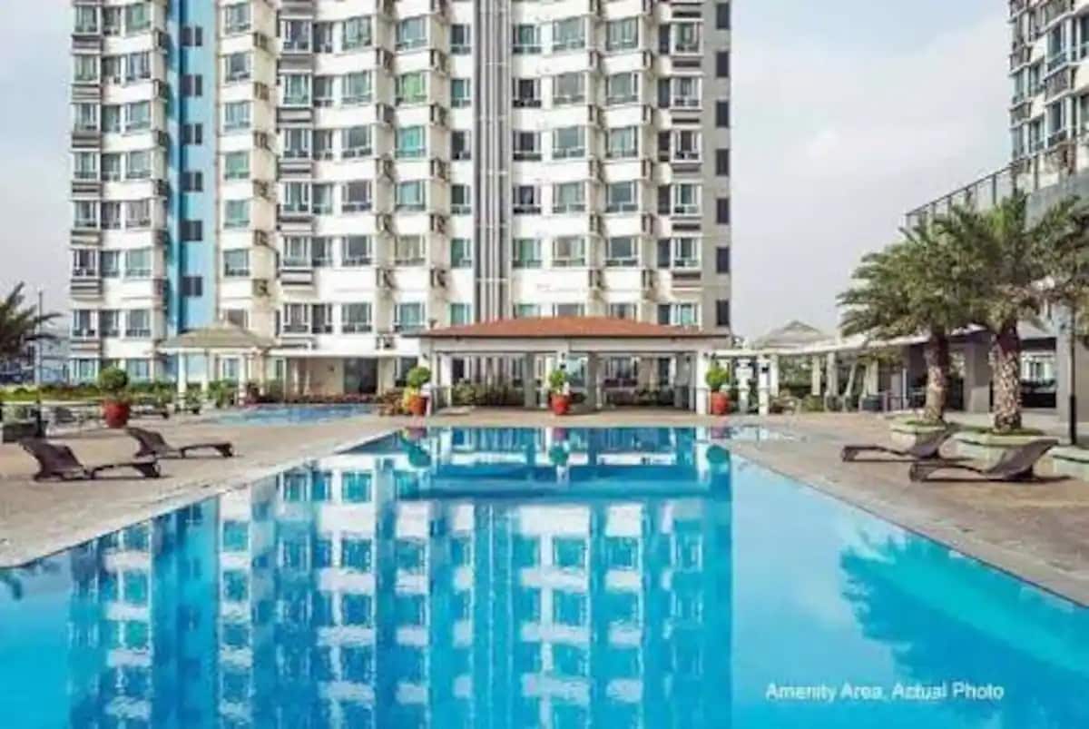 A large swimming pool reflects the surrounding buildings, featuring a paved area with lounge chairs positioned nearby. Lush potted plants and covered seating arrangements enhance the outdoor space, creating an inviting area for relaxation.