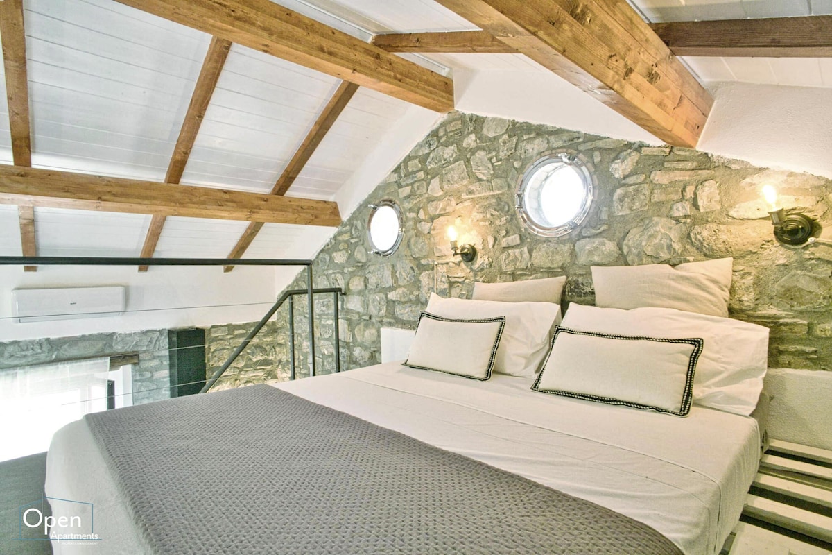 A charming bedroom features a spacious bed adorned with subtle patterned linens and accent pillows. Stone walls complement the modern wooden beams overhead. Unique circular windows add natural light, while soft lighting fixtures enhance the serene ambiance of the space.