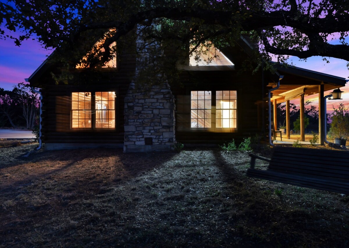 A log cabin exterior is showcased at twilight, featuring large windows that illuminate the interior. A stone chimney is prominently displayed, and a porch swing rests nearby under a shaded tree, inviting relaxation while surrounded by natural scenery.