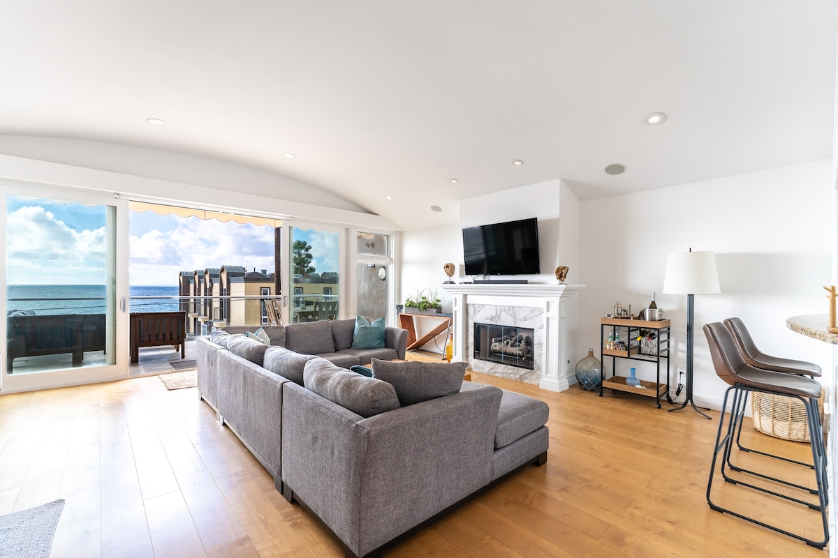A spacious living area features a comfortable gray sectional sofa, facing a white, modern fireplace. Large windows allow for abundant natural light and showcase ocean views. A mounted television is positioned above the fireplace, while a small dining table and bar stools are placed nearby.