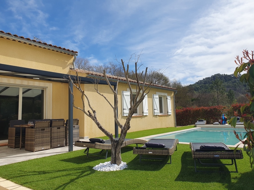 The villa's exterior features a well-maintained garden, complemented by a swimming pool and seating area. Sun loungers are arranged around the pool, and a barren tree stands among the lush grass. The house is illuminated by natural light, with large windows visible.