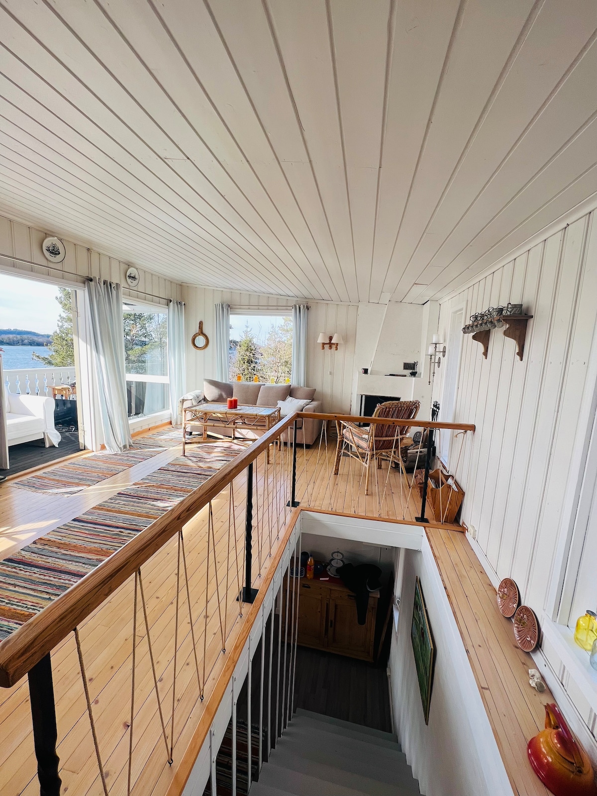 The airy living space features large windows that offer views of the surrounding nature. A wooden staircase leads to the lower level. Light-colored walls and a wooden floor create a welcoming environment, while a comfortable seating area is arranged near a fireplace.
