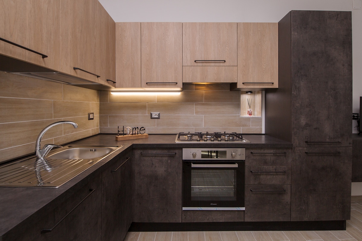A modern kitchen is presented, featuring wooden cabinetry and dark surfaces. A stainless steel sink and a gas stove are visible, alongside an oven integrated into the cabinetry. Soft lighting highlights the work area, creating an efficient cooking environment.