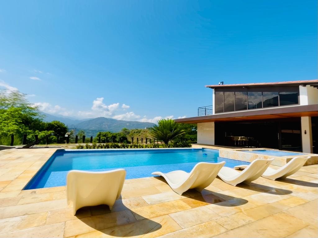 A clear blue pool is surrounded by light-colored stone flooring and sleek white loungers, offering a place to relax. The expansive glass windows of the villa provide a view of the mountains in the background, while greenery adds to the tranquil setting.