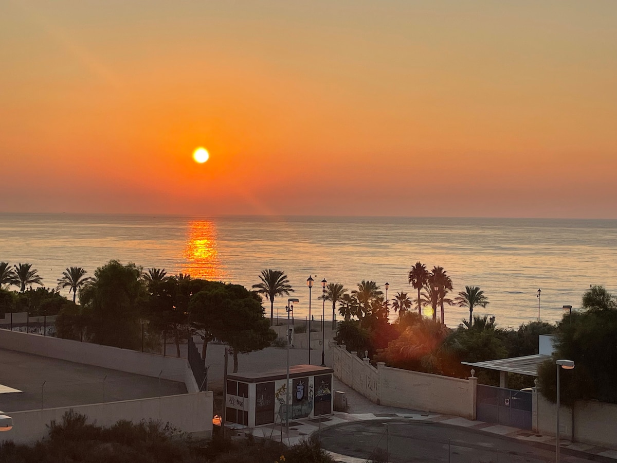 A serene seaside view is captured at sunset, with the sun descending into the horizon, reflected on the calm waters. Silhouettes of palm trees are visible along the shore, while distant structures frame the peaceful landscape.