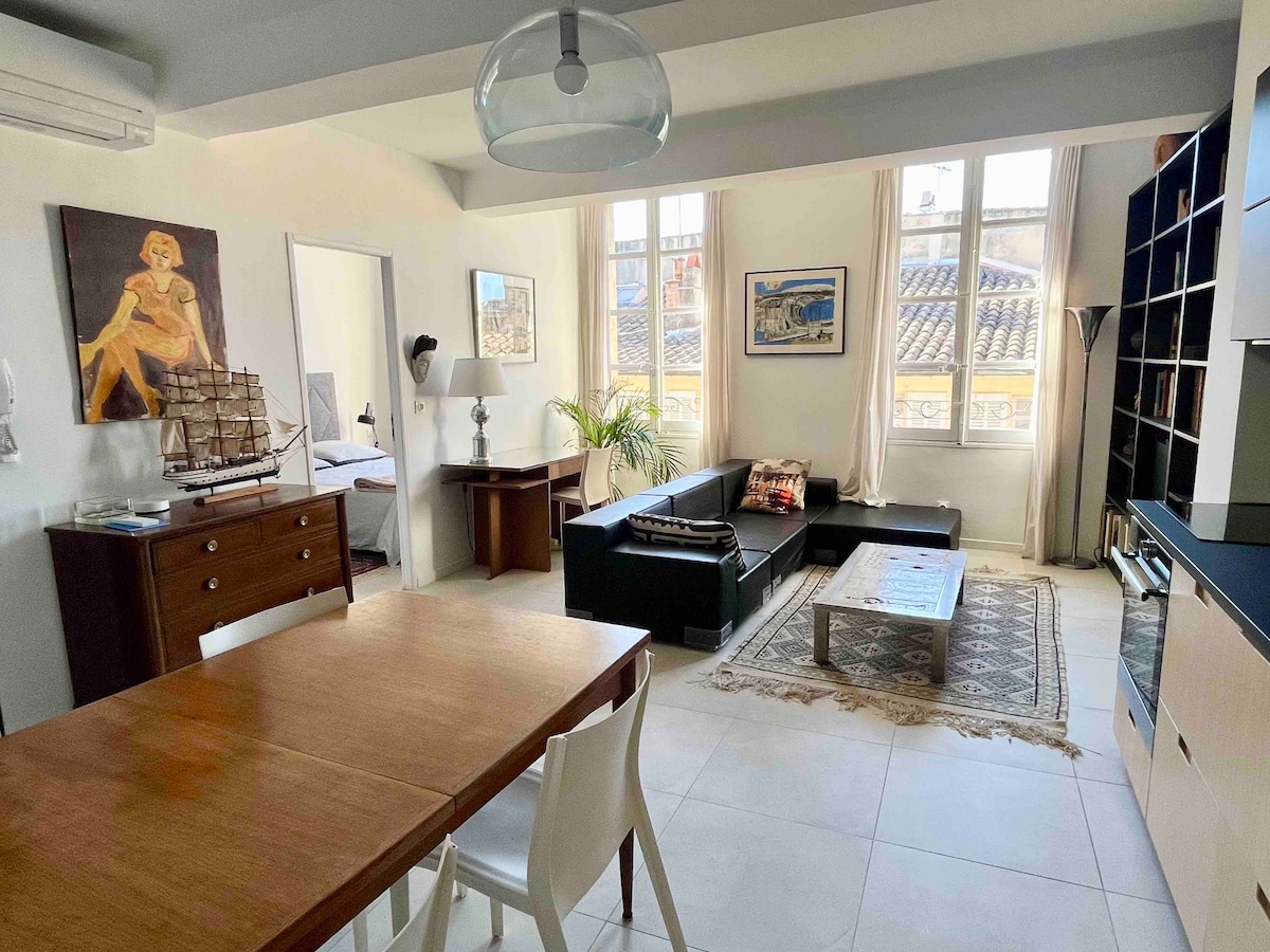 A spacious living area features a dining table made of wood, accompanied by white chairs. A comfortable dark sofa is positioned near large windows with soft curtains. An artwork hangs on the wall, and a coffee table sits on an area rug, enhancing the functional layout.