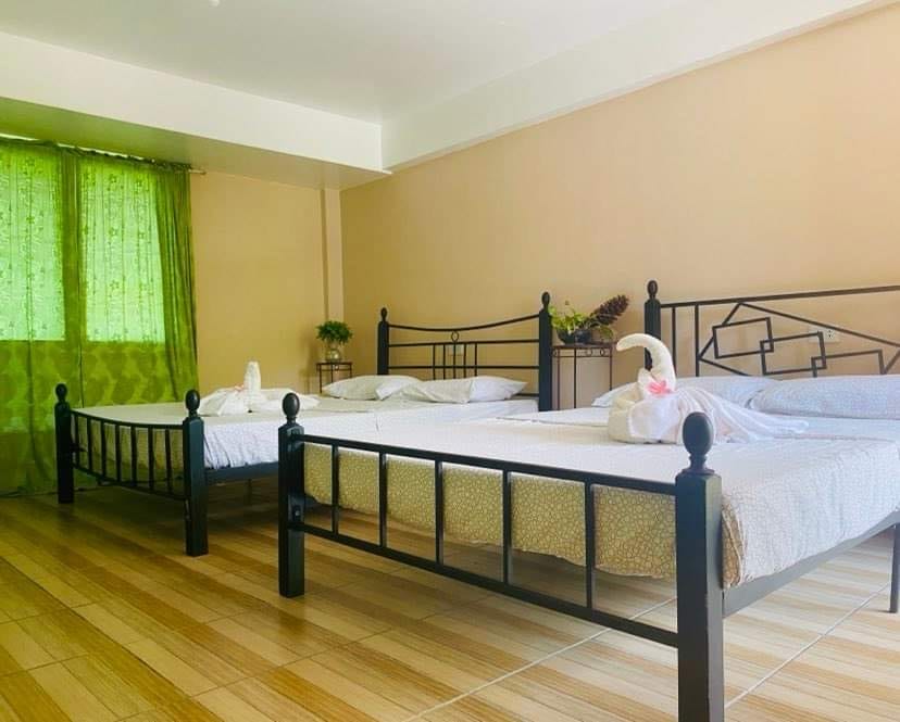 A bright and spacious bedroom features two metal-framed beds, adorned with simple bedding. Natural light filters through green curtains, casting a soft hue across the room. Decorative towel swans are arranged gracefully on each bed, enhancing the inviting atmosphere.