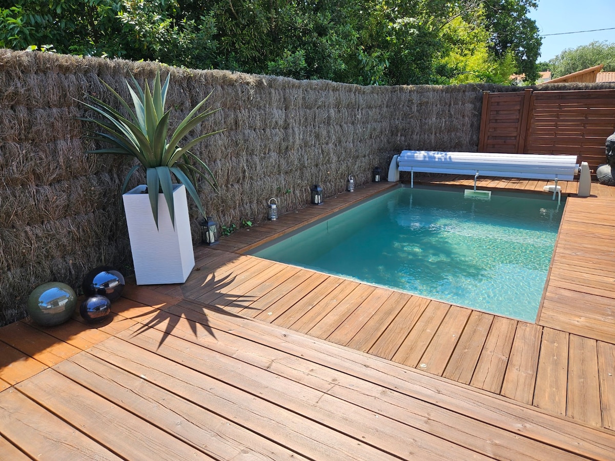 A serene outdoor pool area is surrounded by wooden decking. Lush greenery is visible in the background, and modern decorative elements, including a large potted plant and smooth stones, enhance the space. Subtle lighting fixtures are positioned along the perimeter of the pool.