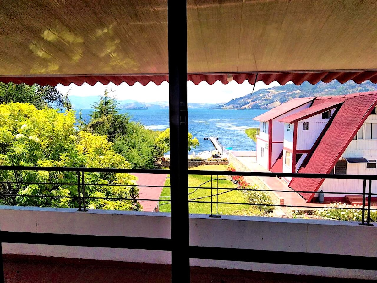 A panoramic view reveals a serene lake stretching towards the horizon, framed by lush greenery. A private dock extends into the water, while charming buildings with red roofs stand nearby, adding to the inviting landscape.