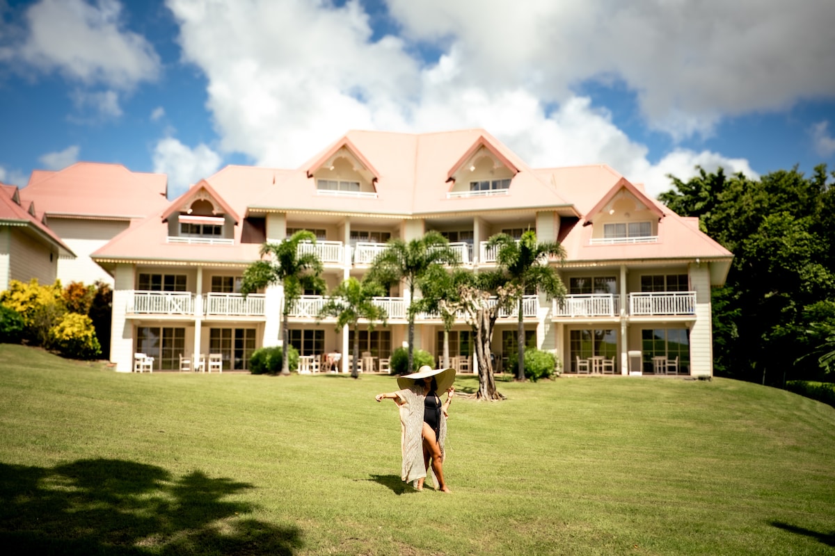 A well-maintained villa complex is depicted, featuring multiple balconies adorned with white railings. Lush green lawns surround the building, complemented by vibrant tropical foliage. A person is seen joyfully standing on the grass, styled in a comfortable outfit, under a partly cloudy blue sky.