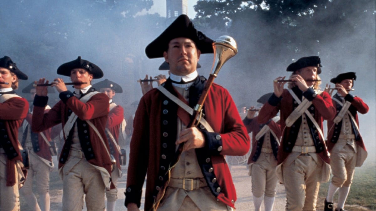 A group of individuals dressed in colonial military uniforms performs with instruments, set against a backdrop of fog and trees. The style reflects 18th-century attire, with red and white colors prominently displayed, while the atmosphere evokes a sense of historical reenactment.