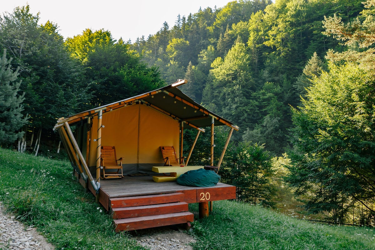 A glamping tent is positioned on a wooden deck, surrounded by lush greenery. The tent features an open front with two wooden lounge chairs and a large cushion. A spacious terrace allows for relaxation while taking in the serene forest views.