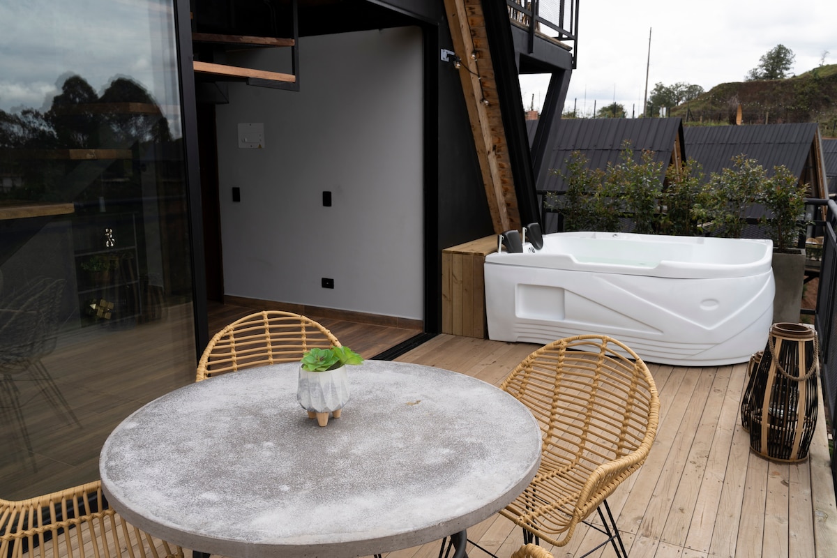 An outdoor balcony area features a round concrete table surrounded by two wicker chairs. A white hot tub is positioned nearby, while wooden deck flooring completes the space, providing a relaxing spot for enjoyment.
