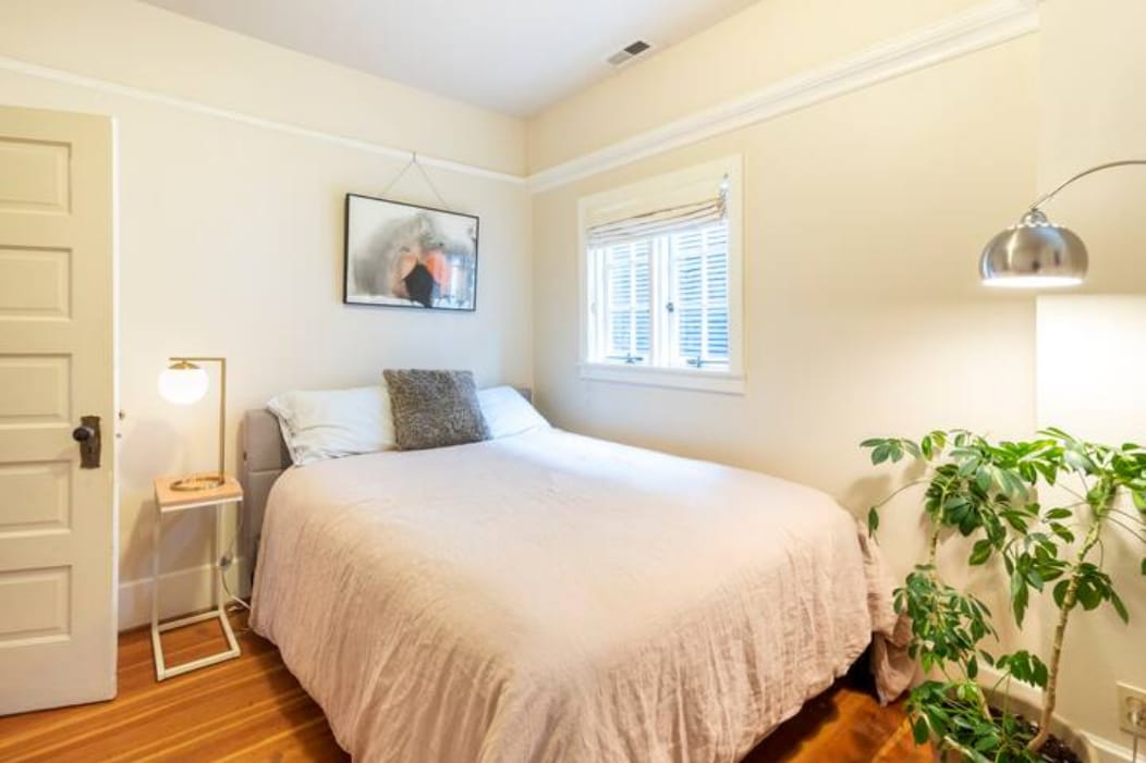 A private bedroom features a spacious bed covered with a light-colored blanket. A piece of art hangs on the wall above the bed. A window allows natural light to fill the room, while a leafy plant adds a touch of greenery beside the bed.