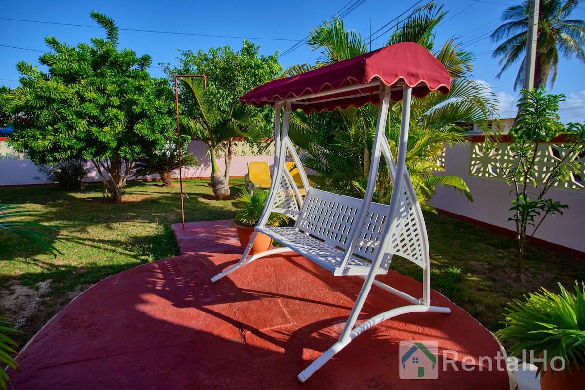 A white swing is positioned on a circular red platform, surrounded by lush greenery and palm trees. The swing features a red canopy and offers a relaxing view of the tranquil garden area, inviting guests to enjoy the serene outdoor atmosphere.