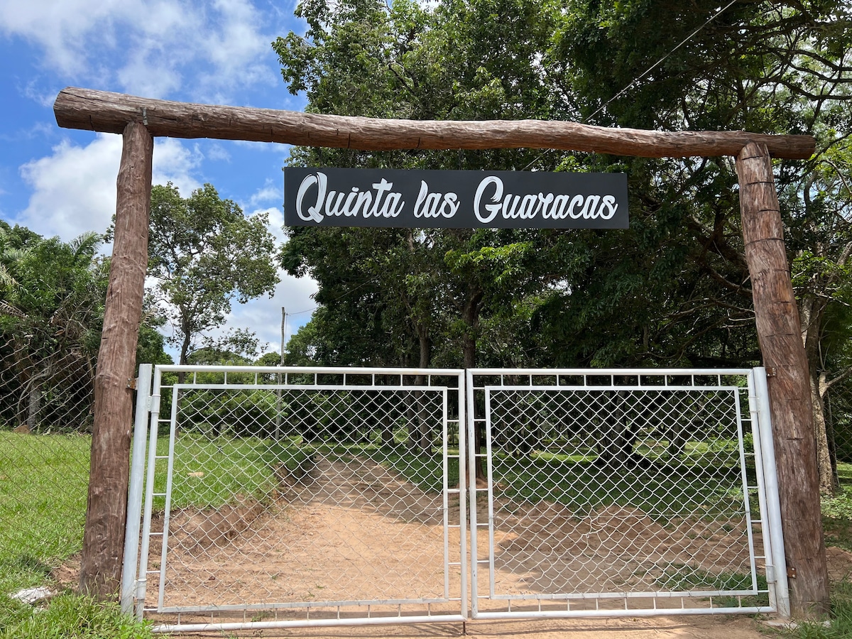 A wooden entrance gate features the name 'Quinta los Guaracas' displayed prominently. The gate is framed by sturdy wooden posts, with a grassy area visible beyond. Lush trees surround the entrance, providing a natural setting with a clear blue sky above.