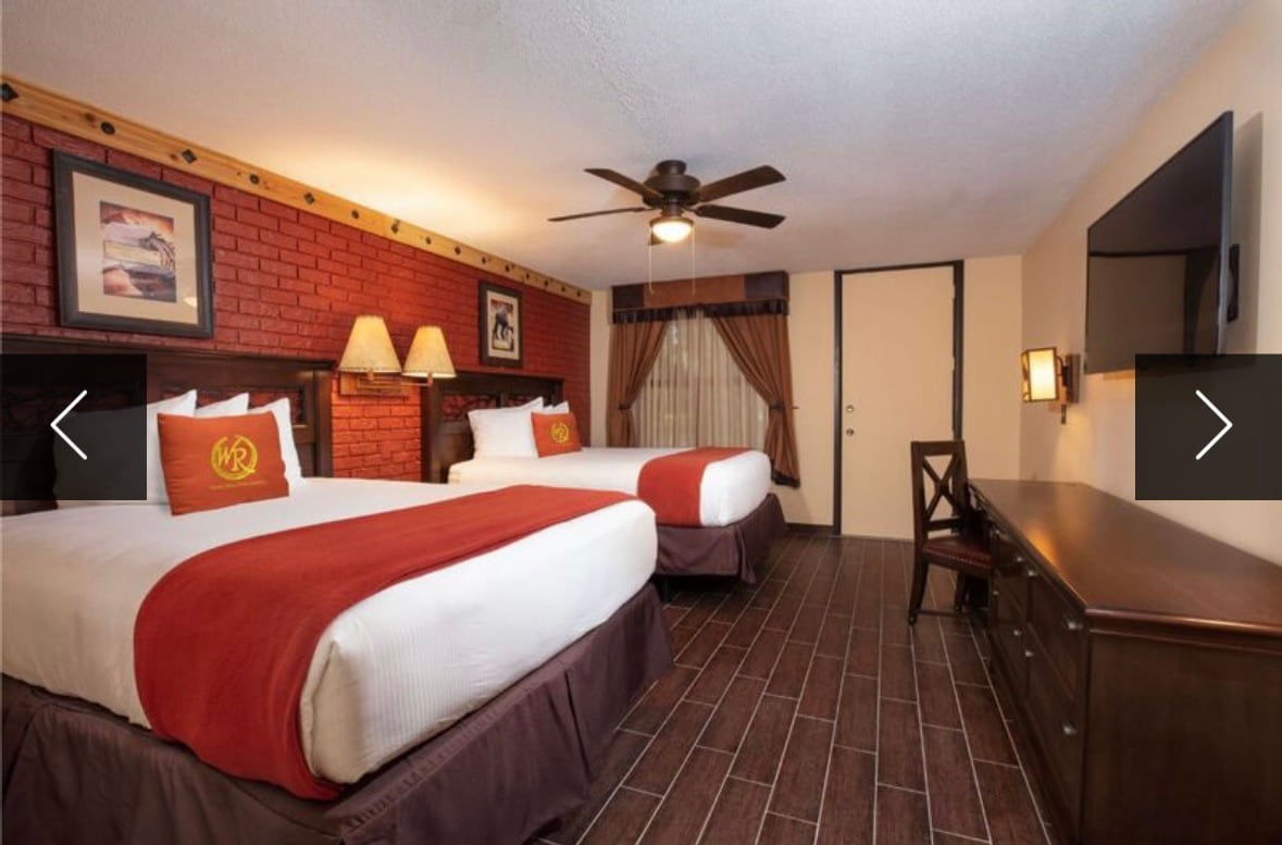 The room features two Queen beds with red and white bedding, bedside lamps, and a brick accent wall. A ceiling fan is mounted above, while a flat-screen TV and a wooden dresser are displayed opposite the beds. Natural light filters in through sheer curtains.
