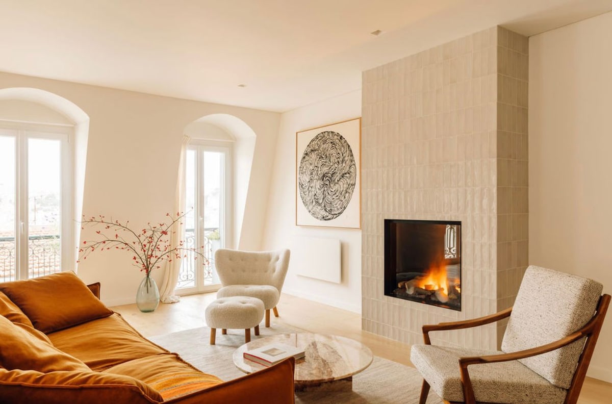 A spacious living area features a warm-toned sofa, a round coffee table, and two accent chairs. Artwork on the wall adds character, while a fireplace serves as a focal point. Large windows allow natural light to illuminate the space, offering a view of the surroundings.