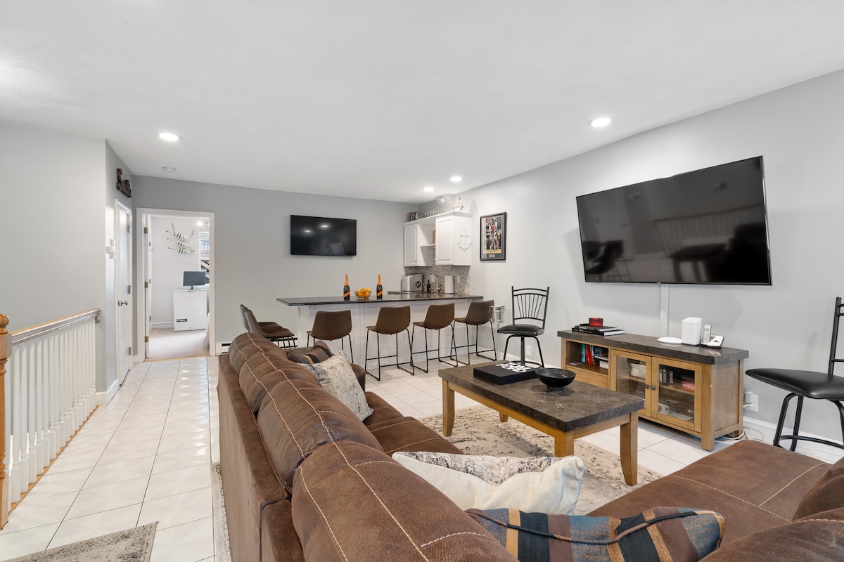 A spacious living area is displayed with a large sectional couch facing two mounted flat-screen televisions. A wooden coffee table occupies the center of the room, complemented by a kitchen bar featuring seating for five. Neutral-colored walls highlight the modern décor.