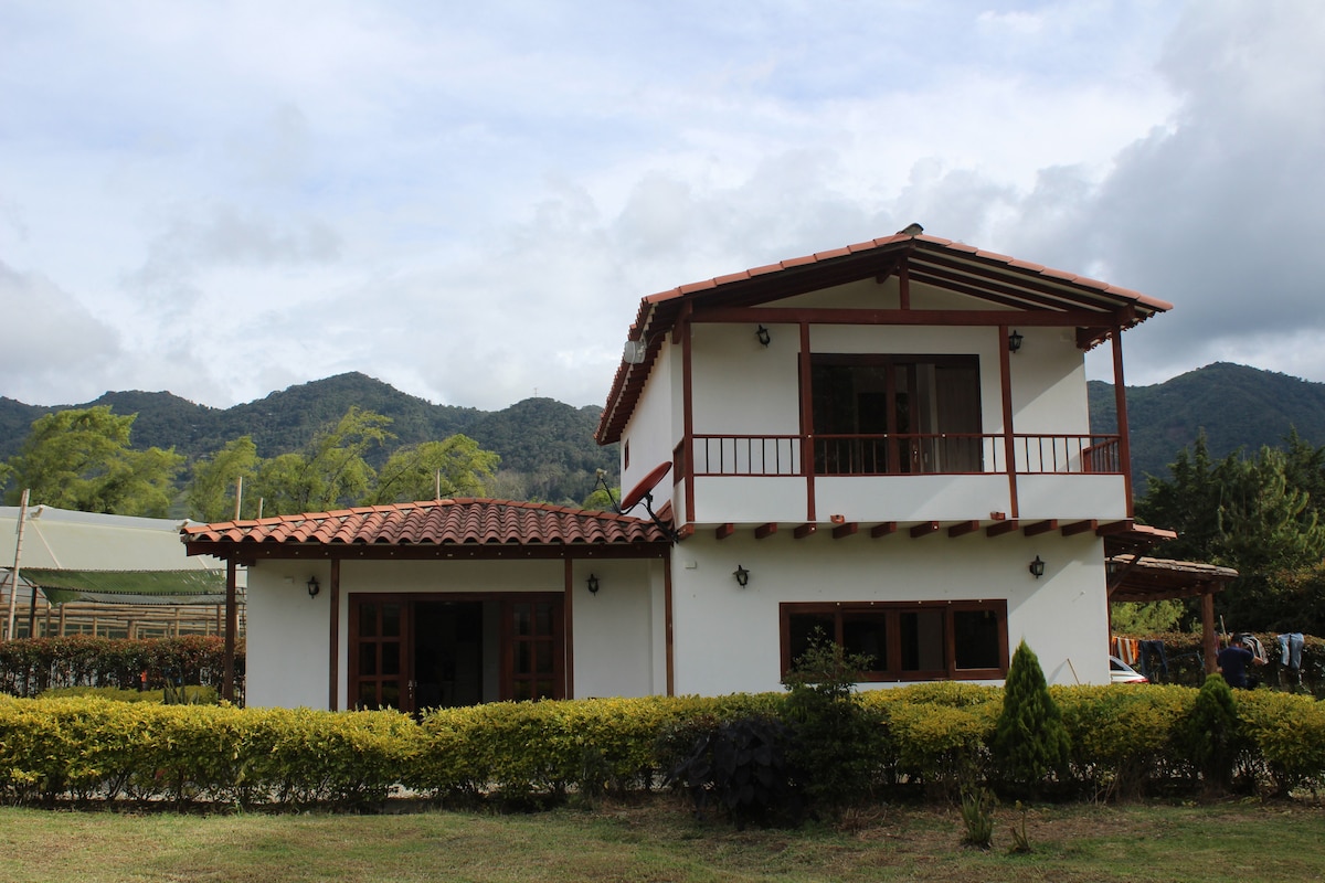 The two-story house features a traditional design with a terracotta roof and white walls. Large windows with wooden frames allow natural light to fill the interior. The surrounding landscape includes rolling hills and greenery, creating a peaceful rural setting.