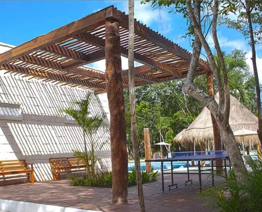 An outdoor area is highlighted by a wooden pergola supported by thick posts. Sunlight casts shadows on the ground, and nearby, a thatched-roof structure and a ping pong table are visible. Lush greenery surrounds the space, providing a serene backdrop.