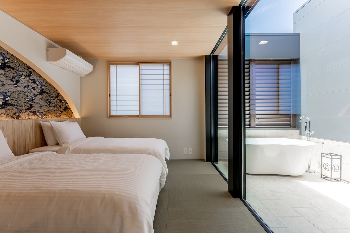 A serene sleeping area features two spacious beds with simple white linens. Natural light flows in through large windows, and a modern outdoor bathtub is visible through glass doors, allowing for seamless outdoor access.
