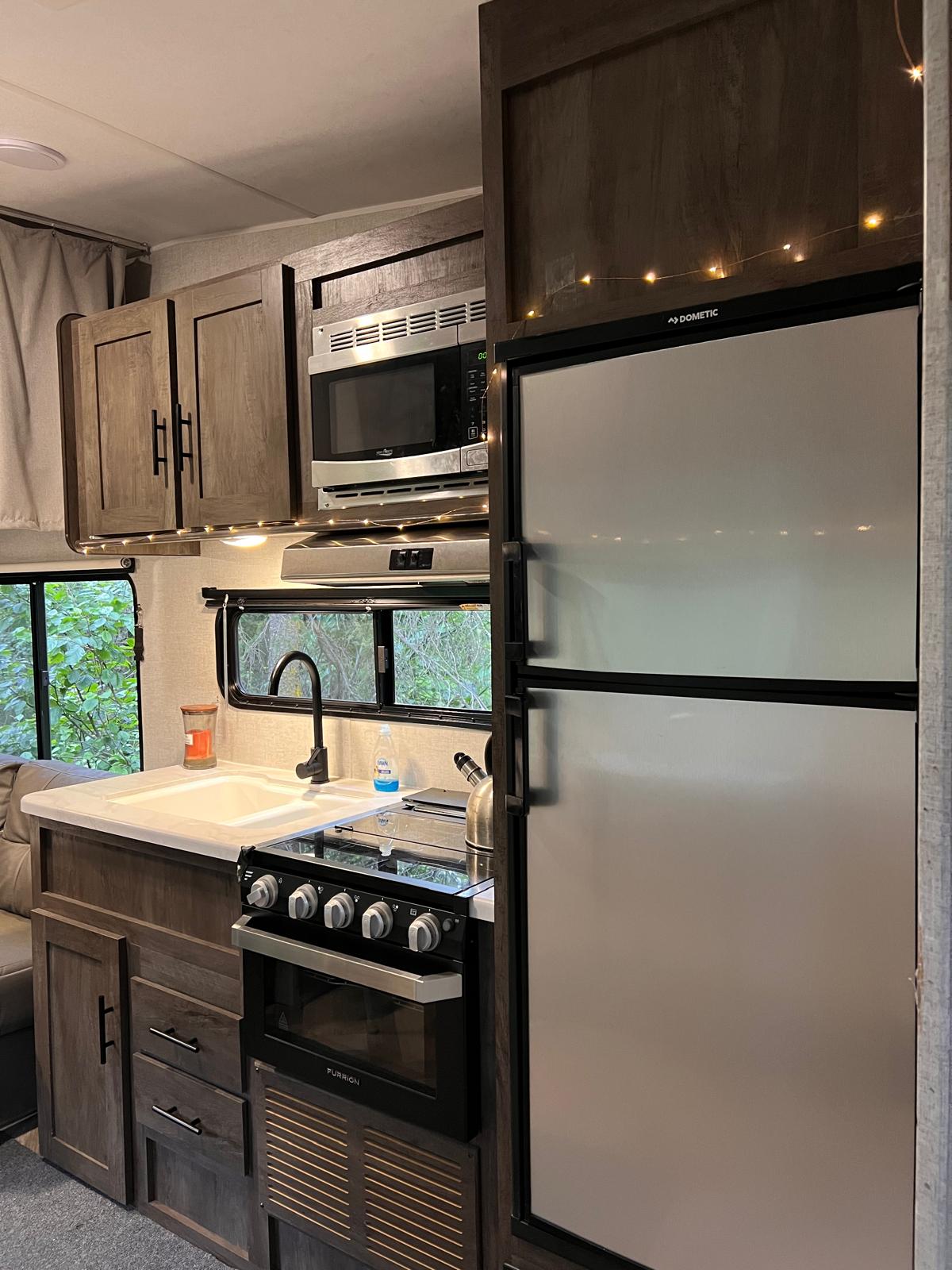 The kitchen area of the travel trailer features dark wood cabinetry and stainless steel appliances, including a microwave, stove, and a refrigerator. A white sink with a modern faucet is visible, surrounded by counter space, with a cozy atmosphere accentuated by soft lighting.