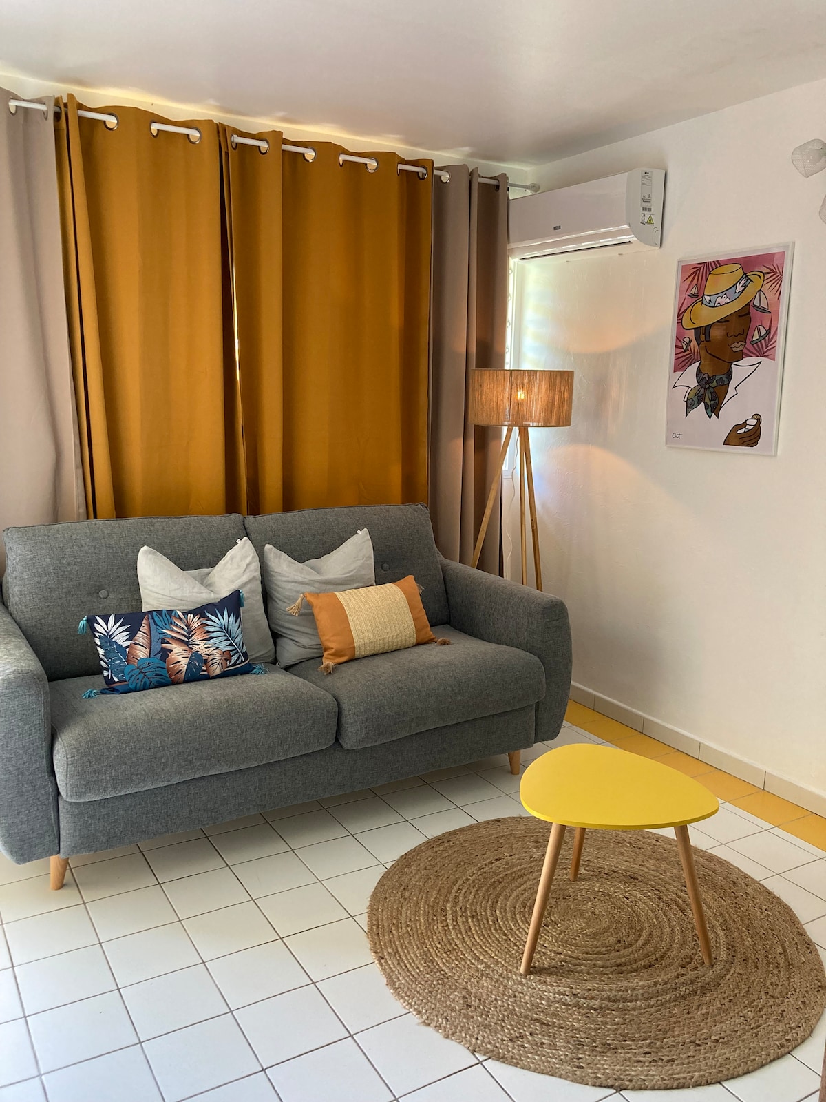 A cozy seating area is featured with a gray sofa adorned with decorative pillows in various colors. A circular woven rug sits under a small yellow table, while large, warm-toned curtains frame a window. A modern floor lamp and wall art complete the inviting space.