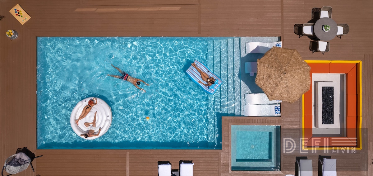 An overhead view of a spacious outdoor area featuring a sparkling pool and a jacuzzi. Several loungers are positioned around the pool, while individuals enjoy relaxation on inflatable floats. An umbrella provides shade over a dining table in the corner, enhancing the inviting atmosphere.