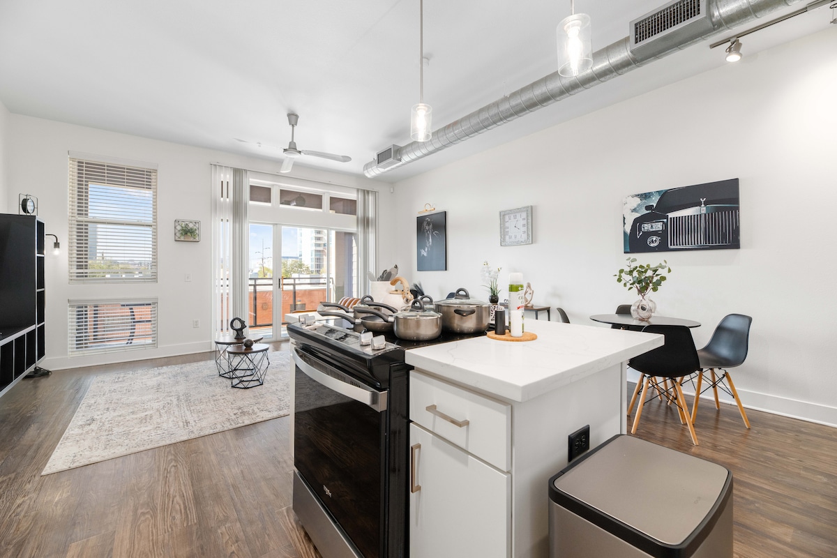 A contemporary kitchen with sleek black appliances is showcased, featuring a stove and numerous cooking pots on the stovetop. A small dining area is visible, complemented by stylish wooden chairs and a circular table. Large windows allow natural light to brighten the open living space.