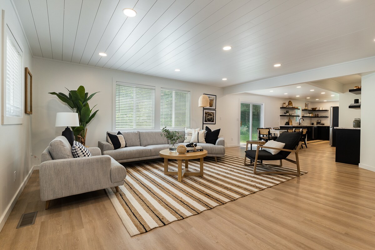 A spacious living area features a light-colored sectional sofa with black and white accent pillows. A round coffee table sits atop a striped rug, while large windows allow natural light to brighten the space. A tall plant adds a touch of greenery in the corner.