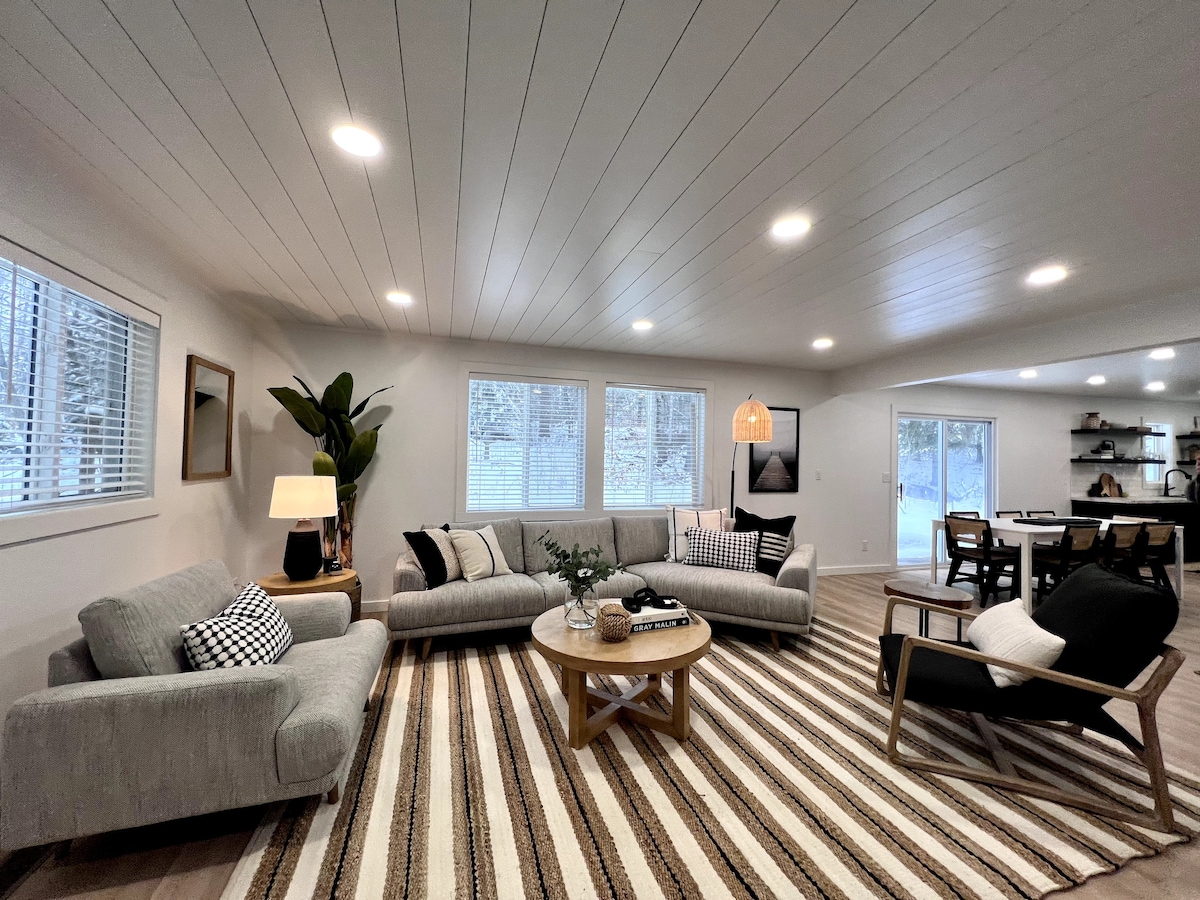 A spacious living area features a gray sectional sofa and a black accent chair, arranged around a wooden coffee table on a striped rug. Bright natural light enters through three windows, highlighting the modern decor and dining area in the background.