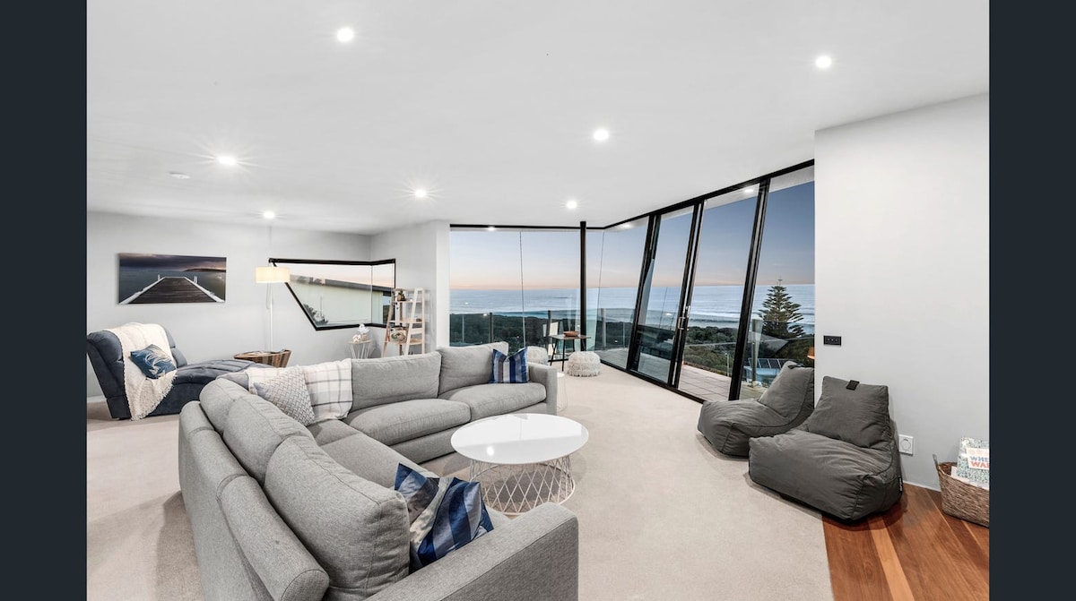 A spacious living area features large floor-to-ceiling windows offering expansive ocean views. Comfortable seating arrangements include a sectional grey sofa and bean bags, complemented by a round coffee table. Soft lighting enhances the bright and airy atmosphere, while a piece of wall art adds a touch of sophistication.